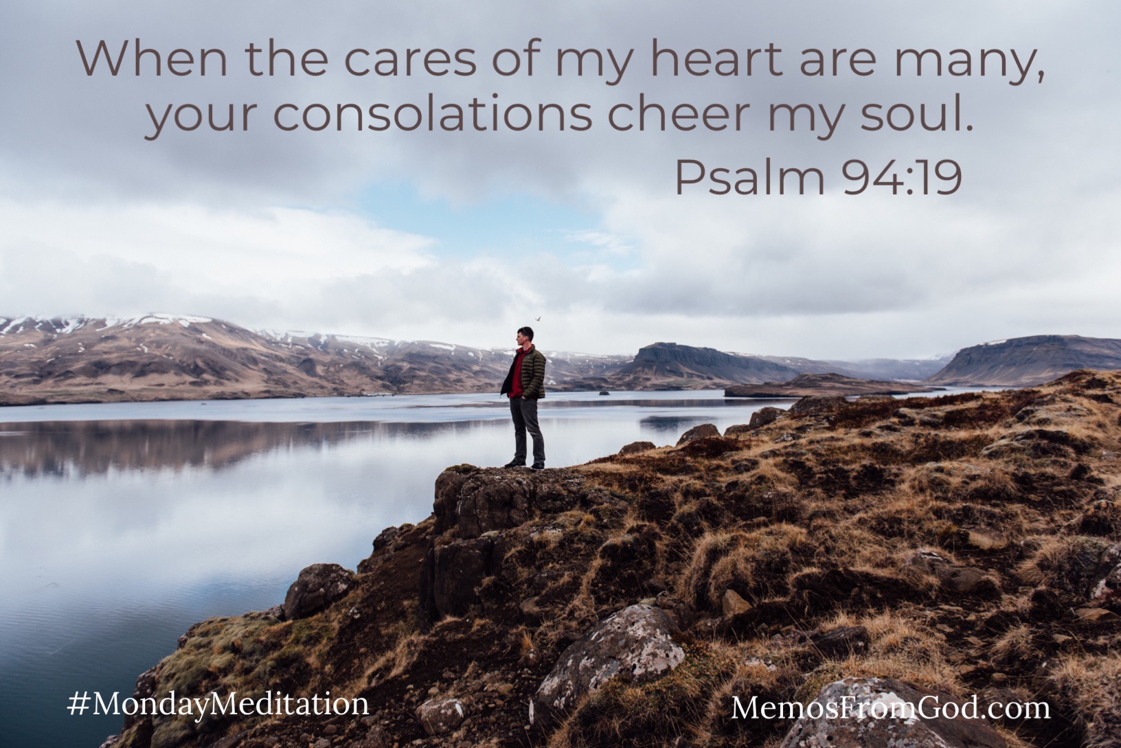 A man standing on a rocky shore looking out at the still water of a lake with a mountain in the background. Caption: When the cares of my heart are many, your consolations cheer my soul. Psalm 94:19